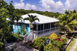 A two-story house with a white roof, surrounded by lush greenery and palm trees.