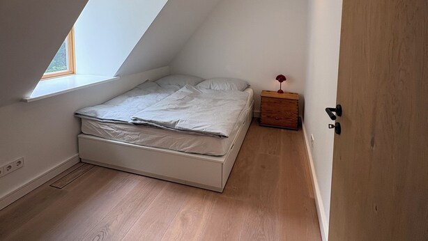 Ein Schlafzimmer mit schrägem Dach, einem Bett, einem kleinen Nachttisch und einem Fenster.