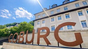 Een groot metalen bord met de tekst 'COBURG' voor een gebouw met meerdere ramen.
