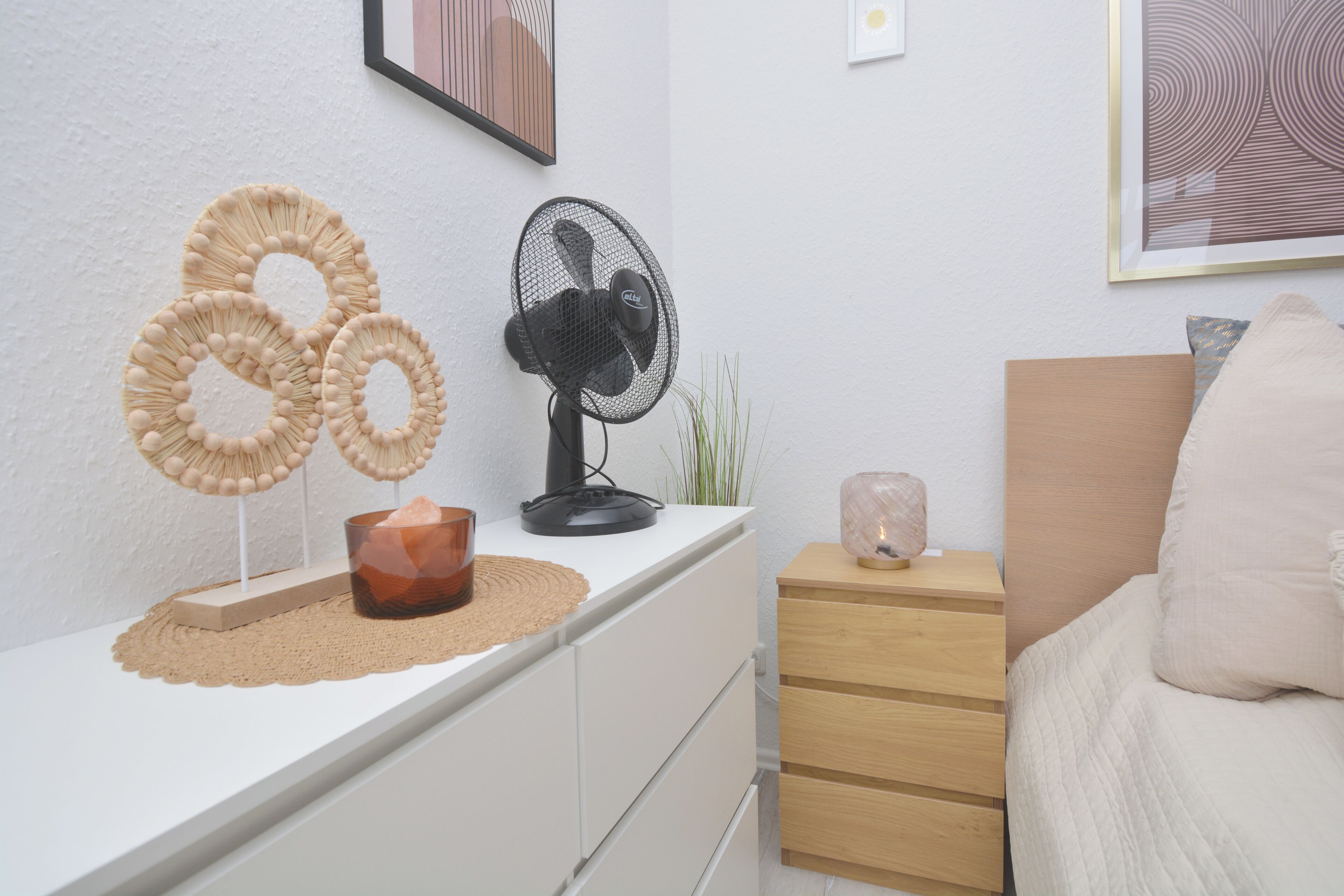 Ein Schlafzimmer mit einem weißen Schminktisch, einem Nachttisch mit Lampe, einem Bett mit weißem Kissen und Wandbildern.