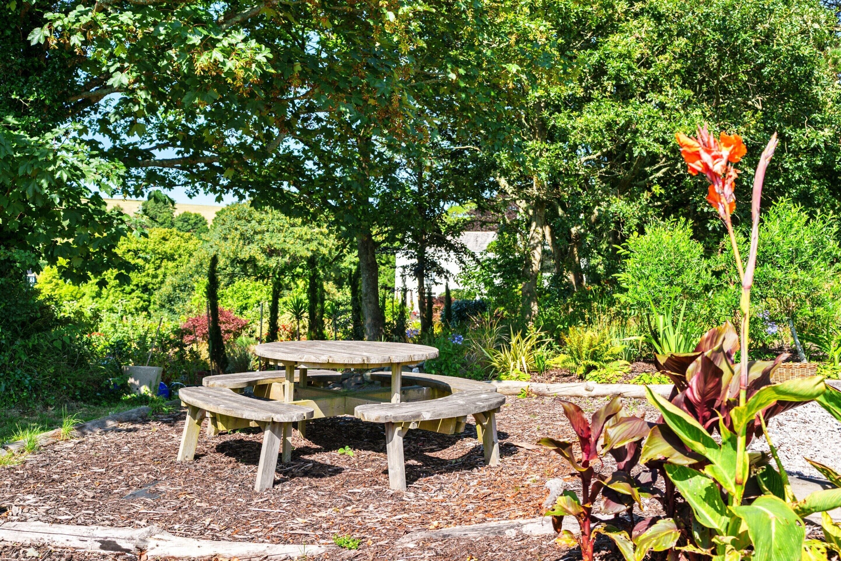 A wooden picnic table surrounded by mulch, with a garden in the background.