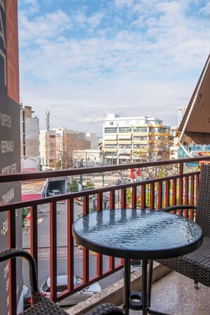 Un balcon avec une table ronde et des chaises, donnant sur une rue bordée d’immeubles et d’un lave-auto.