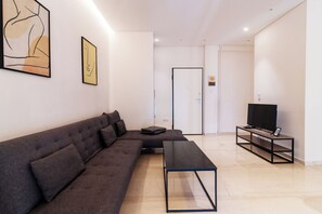 A modern living room with a sectional sofa, a coffee table, a TV on a stand, and artwork on the walls. - Cozy - Bright & New Apartment - Downtown (Athina)