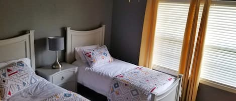 A bedroom with two beds, a nightstand with a lamp, and a window with blinds.