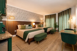 Habitación de hotel con una cama, dos otomanos verdes, una mesita de noche de madera con una lámpara, una mesa de comedor con sillas y un papel tapiz estampado.