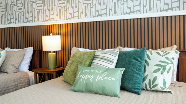 Ein ordentlich bezogenes Bett mit vielen Kissen, einer Nachttischlampe und einer Wand mit geometrischem Muster.