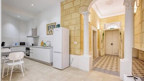 A modern kitchen with white appliances, a dining area with a white chair, and a hallway with a patterned floor. - Charming 1BR Maltese Maisonette w/ Terrace, Luqa (Ħal Luqa)