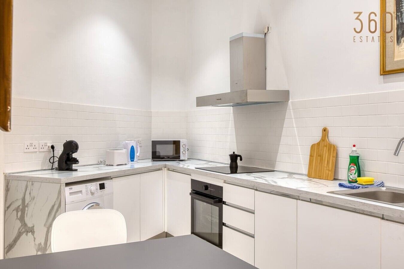 Una cocina moderna con armarios blancos, microondas integrado, cafetera y lavabo con tabla de cortar y jabón para lavar platos.