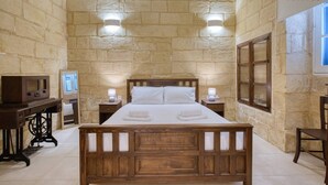 A bedroom with a wooden bed, two bedside tables with lamps, a desk with a computer, and a stone wall. - Charming 1BR Maltese Maisonette w/ Terrace, Luqa (Ħal Luqa)