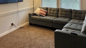 A living room with a sectional sofa, a flat-screen TV, a ceiling fan, and a window with blinds. - Cozy 2-bedroom house in delightful San Antonio with AC (San Antonio)