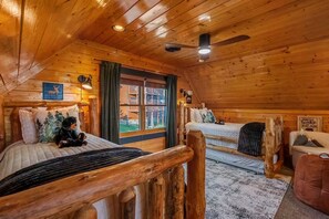A log cabin bedroom with two beds, a ceiling fan, and a window with a view of a wooden deck. - Panoramic Views|Hot Tub|Game Room|Resort Pool (Sevierville)