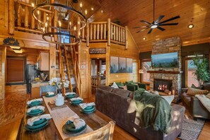 A spacious log cabin interior with a dining area, a fireplace, a living room with a sofa, and a staircase leading to an upper level. - Panoramic Views|Hot Tub|Game Room|Resort Pool (Sevierville)
