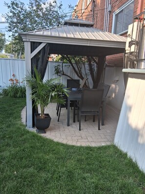 A covered patio area with a table and chairs, surrounded by a white fence and potted plants. - Elegant and cozy  5 1/2 appartement with pool (Longueuil)