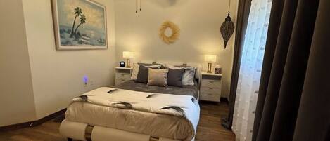 A bedroom with a bed, a ceiling fan, a nightstand, a wall-mounted lamp, and a painting.