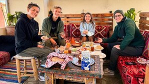 Un groupe de personnes est assis autour d’une table, devant de la nourriture et des boissons, près d’un banc en bois et d’un tapis à motifs.