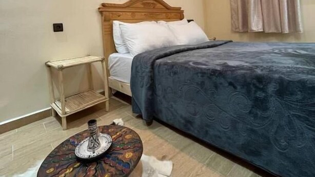 Un dormitorio con cama, una mesita redonda con un adorno, estante en la pared y una ventana con cortinas.