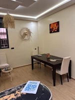 Un comedor con mesa de madera oscura, sillas blancas, lámpara colgante y una obra de arte en la pared.
