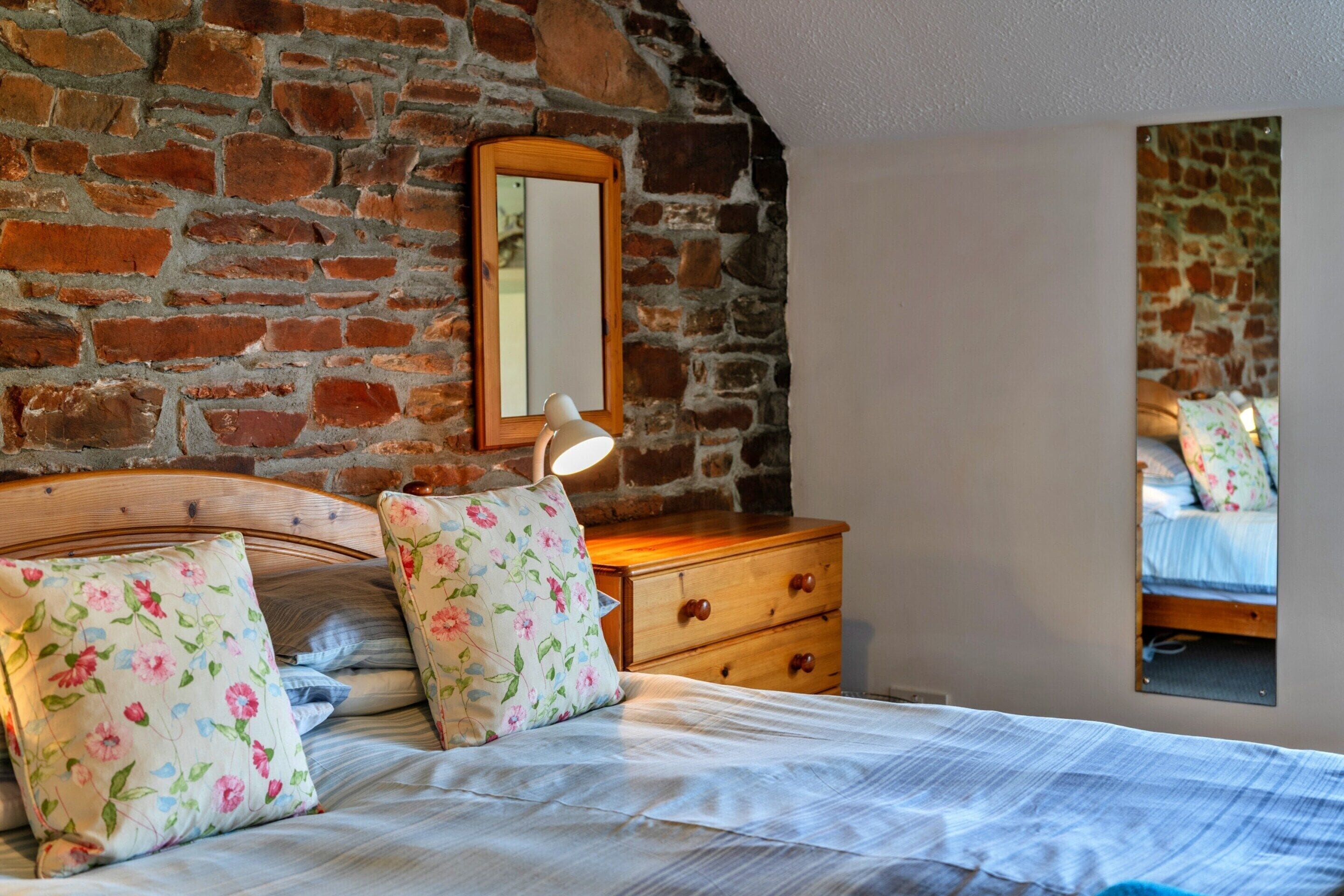 A bedroom with a brick wall, a wooden headboard, a bedside lamp, a dresser with floral-patterned drawers, and a bed with floral pillows.