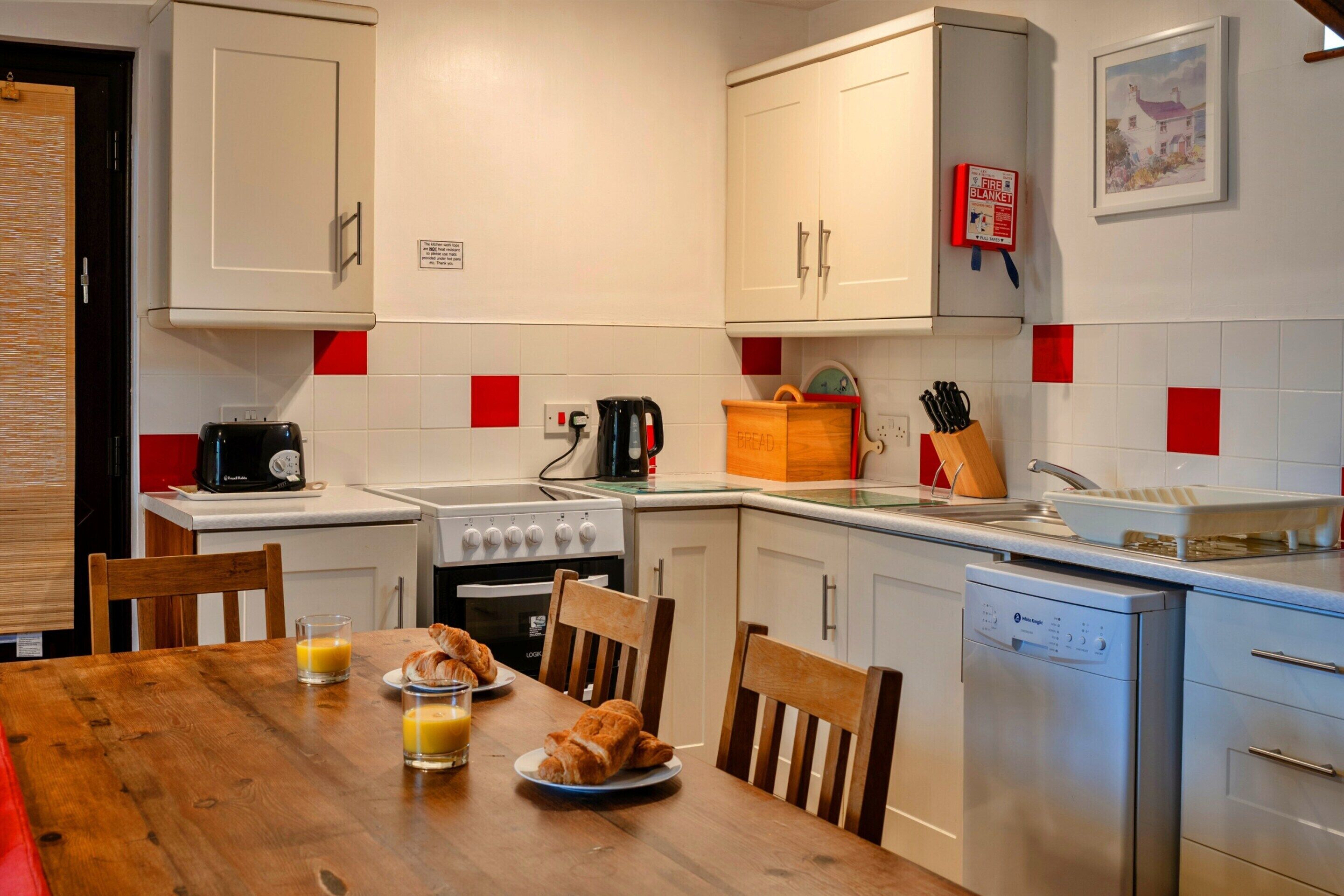 A modern kitchen with white cabinets, a stove, an oven, and a dishwasher. There is a dining table with chairs, a coffee maker, and a bread box.