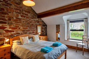 A bedroom with a brick wall, a wooden bed, a nightstand with a lamp, a dresser, a window with curtains, and a chair.