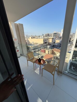 Un balcó amb dues cadires de vimetla i una taula petita, amb vista a un paisatge urbà ple d'edificis i un cel serè.