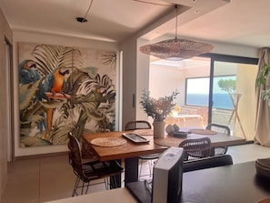 A dining area with a large tropical mural, a table with chairs, and a view of the ocean through a glass door. - Beautiful apartment with a large terrace overlooking the sea (Bandol)