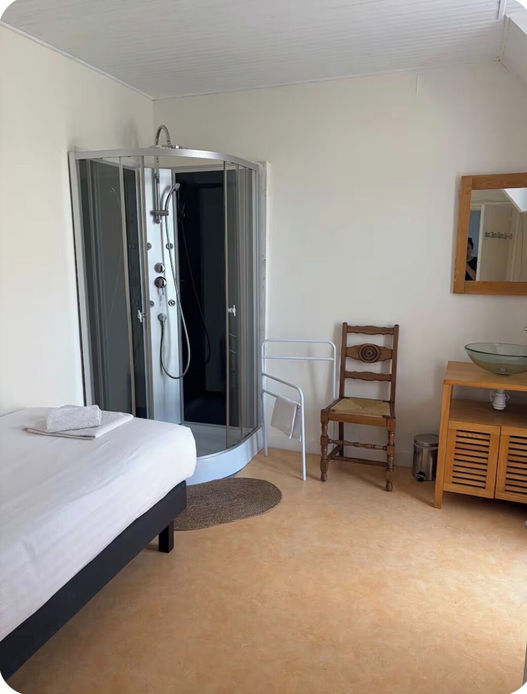 A hotel room with a bed, a wooden chair, a small table with a bowl, and a shower area.