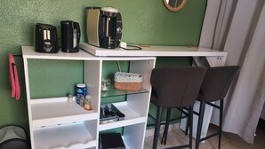 Et tekøkken med kaffemaskine, brødrister og elkedel, en skraldespand, en lille hylde med en dåse og en flaske, samt to barstole.
