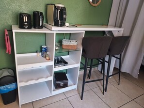 Eine Küchenzeile mit Kaffeemaschine, Toaster und Wasserkocher, einem Mülleimer, einer kleinen Ablage mit einer Dose und einer Flasche sowie zwei Barhocker.
