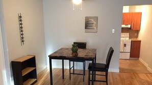 Et spiseområde med et bord og stole, en lille reol, en vase, en loftlampe og et køkken i baggrunden.