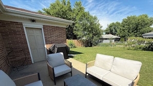 A patio with a brick wall, a white door, a covered grill, a white sofa, a wicker chair, and a small table. - The Brick Haven in Brookings - spacious home in a central location! (Brookings)