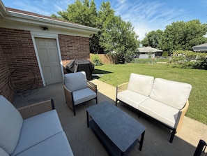 A patio with a brick wall, a white door, a covered grill, a white sofa, a wicker chair, and a small table. - The Brick Haven in Brookings - spacious home in a central location! (Brookings)