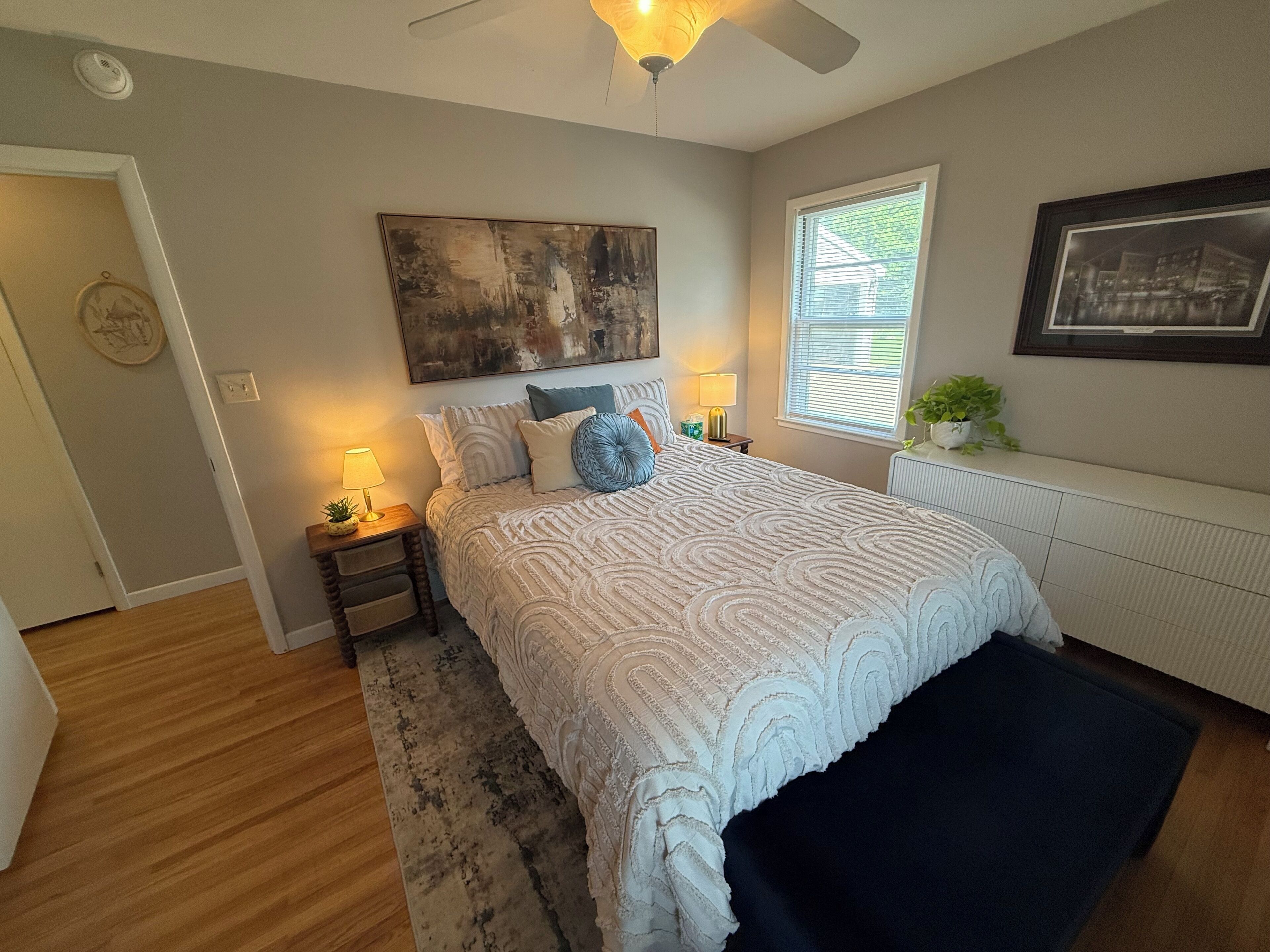 A bedroom with a bed, bedside lamps, a ceiling fan, a window with blinds, and a radiator.