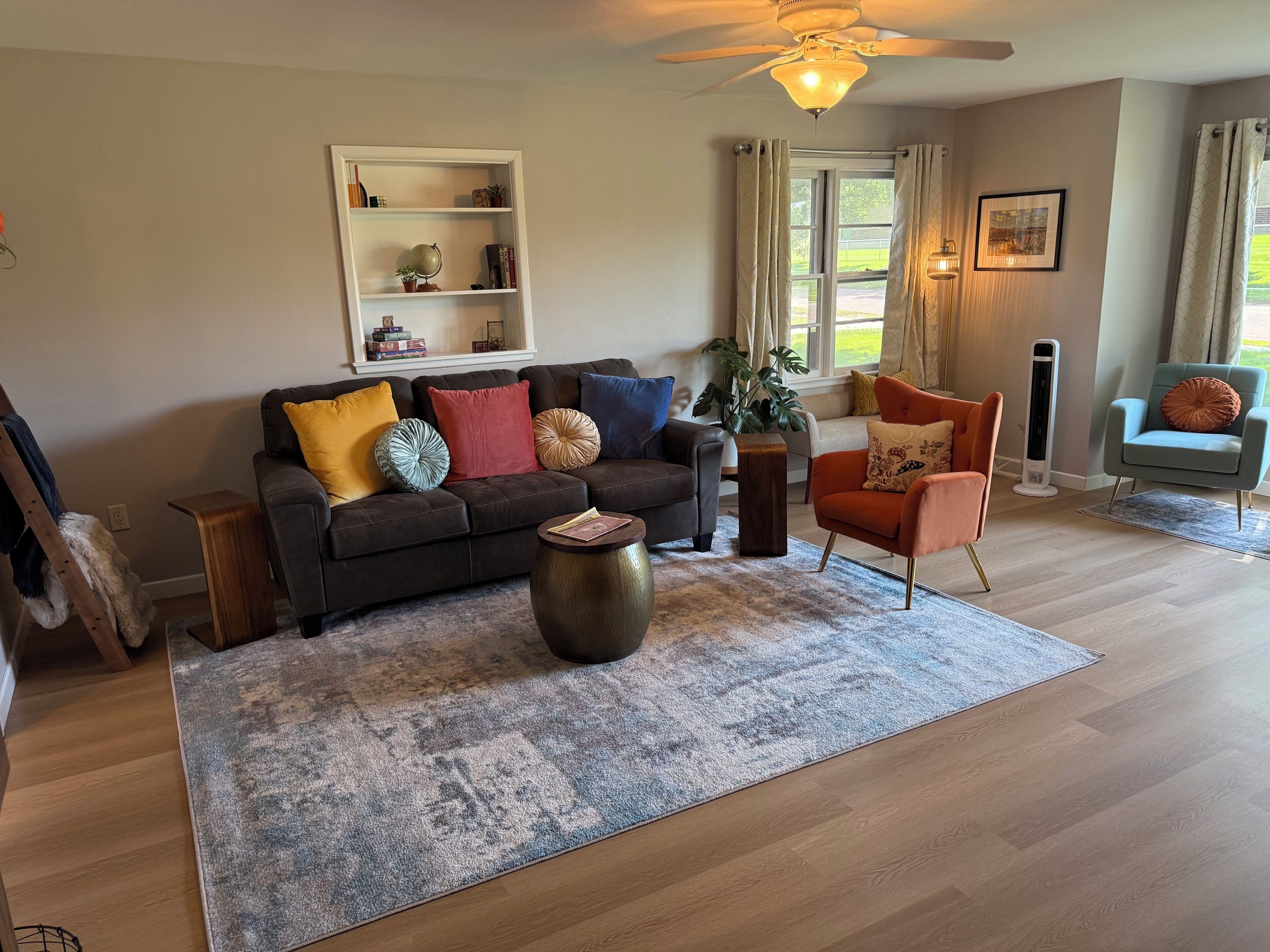 A living room with a brown leather sofa, a coffee table, and a blue armchair. There is a bookshelf with decorative items and a window with curtains.