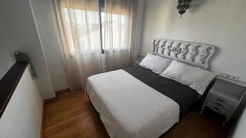 A bedroom with a bed, a nightstand, a wall-mounted lamp, and a window with sheer curtains.