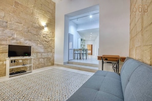 A living room with a stone wall, a flat-screen TV on a white stand, a grey sofa, and a patterned rug. - Stylish 2BR Maltese Maisonette w/ Terrace, Luqa (Ħal Luqa)