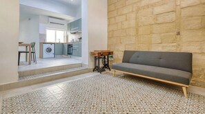 A living room with a grey sofa, patterned floor tiles, a small wooden table, and a kitchen in the background. - Stylish 2BR Maltese Maisonette w/ Terrace, Luqa (Ħal Luqa)