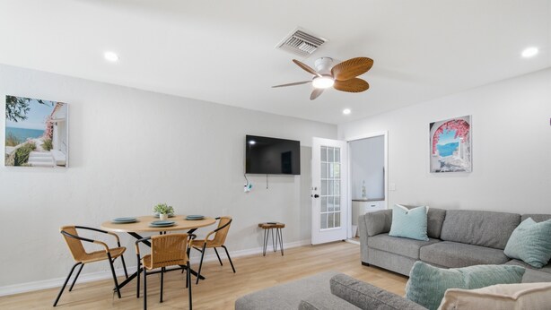 Una sala de estar moderna con un sofá gris, una mesa de comedor con sillas, un ventilador de techo y un televisor de pantalla plana montado en la pared.