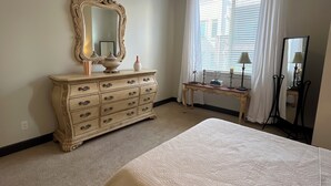 Une chambre à coucher comprenant un lit, une commode, un miroir, un ventilateur de plafond et une fenêtre avec des rideaux.