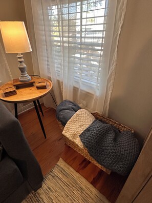 Ein gemütliches Zimmer mit einem runden Holztisch, einer Lampe, einem Korbstuhl und einem kleinen Bett mit Strickdecke.