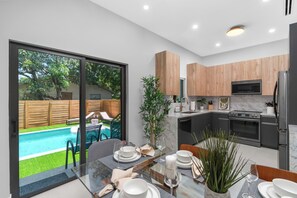 A modern kitchen with wooden cabinets, a built-in oven, and a dining area with a glass table overlooking a swimming pool. - LUXE 4BR Pool Oasis Near Wynwood & Design District (Miami)