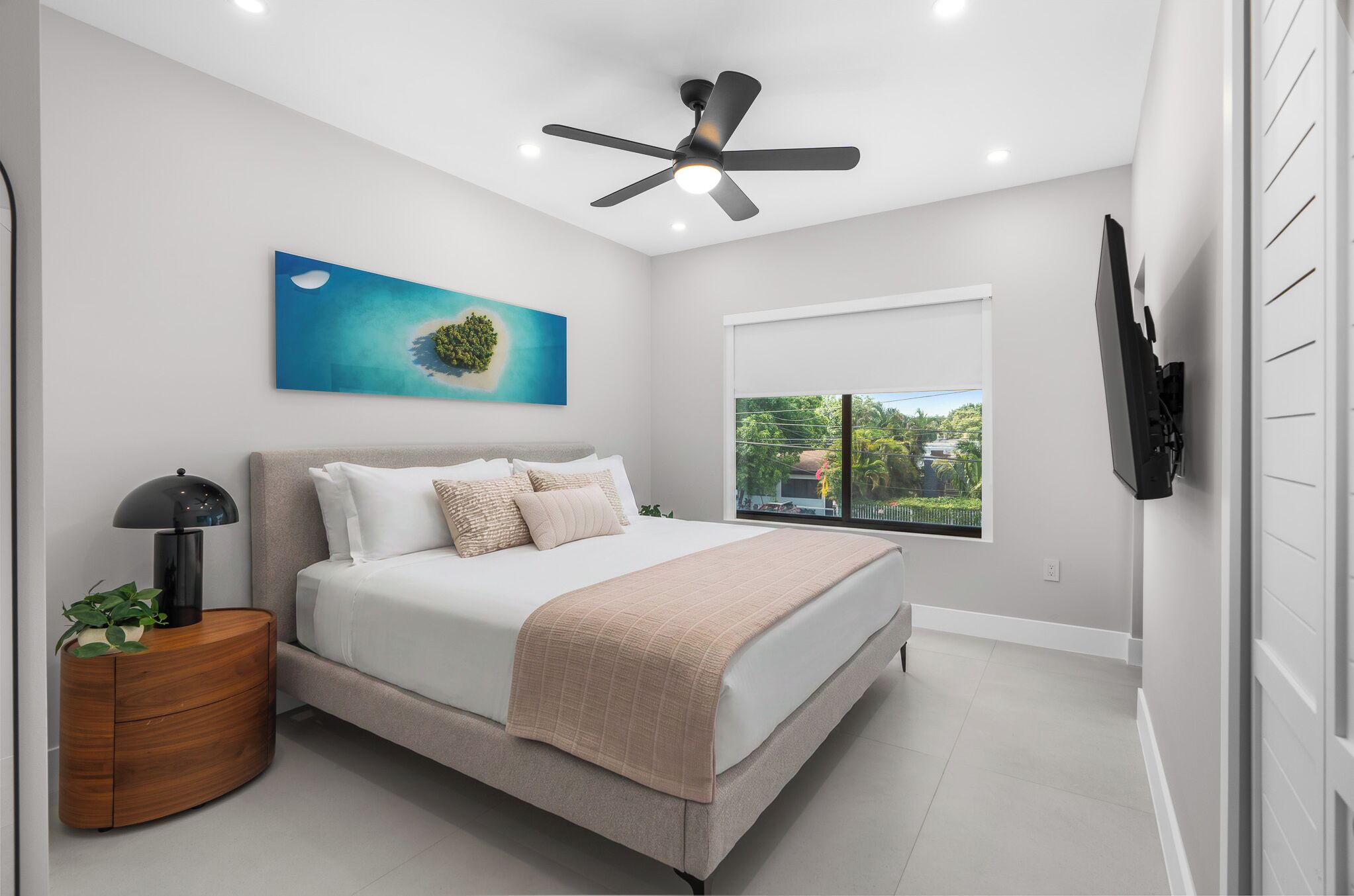 A modern bedroom with a bed, a ceiling fan, a window with a view, and a wall-mounted TV.