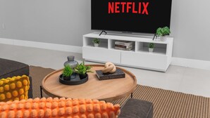 Ein Wohnzimmer mit einem Flachbildfernseher, der 'NETFLIX' zeigt, einer weißen Unterhaltungsanlage mit Büchern und Pflanzen, einem runden Couchtisch mit Dekoration und einem grauen Sofa.