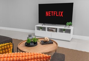 A living room with a flat-screen TV displaying 'NETFLIX,' a white entertainment unit with books and plants, a round coffee table with decorative items, and a gray sofa. - LUXE 4BR Pool Oasis Near Wynwood & Design District (Miami)