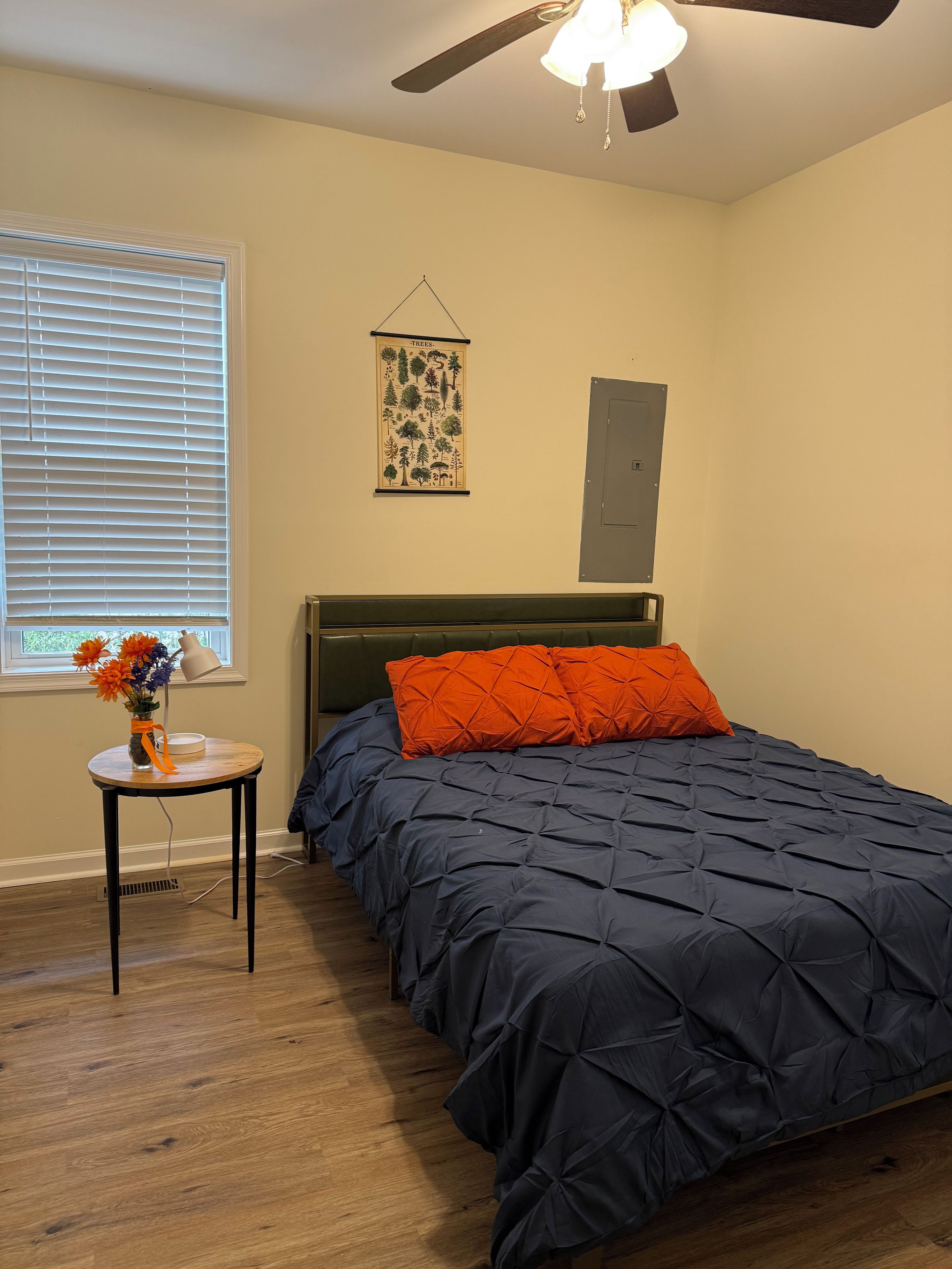 A bedroom with a bed, a wall-mounted light fixture, a window with blinds, and a small table with a vase.
