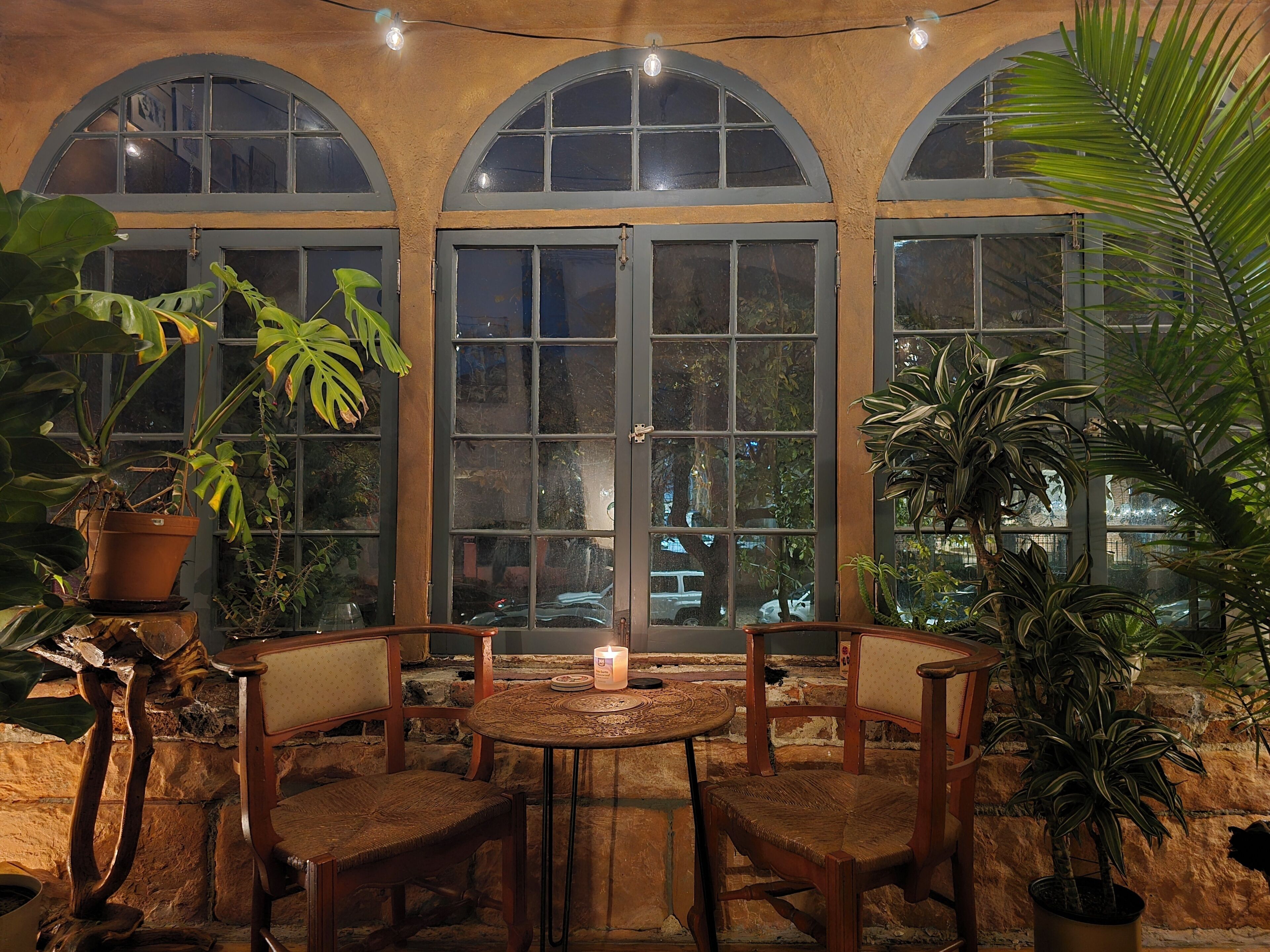Un rincón interior acogedor con una mesa redonda y sillas, rodeado de ventanas amplias y plantas en macetas.