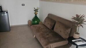 Een woonkamer met een bruine bank, een groene vaas met bloemen, een klein tafeltje met een plant en een klok aan de muur.