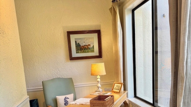 Una habitación de esquina con ventana, una silla, una mesa con lámpara y un cuadro en la pared.