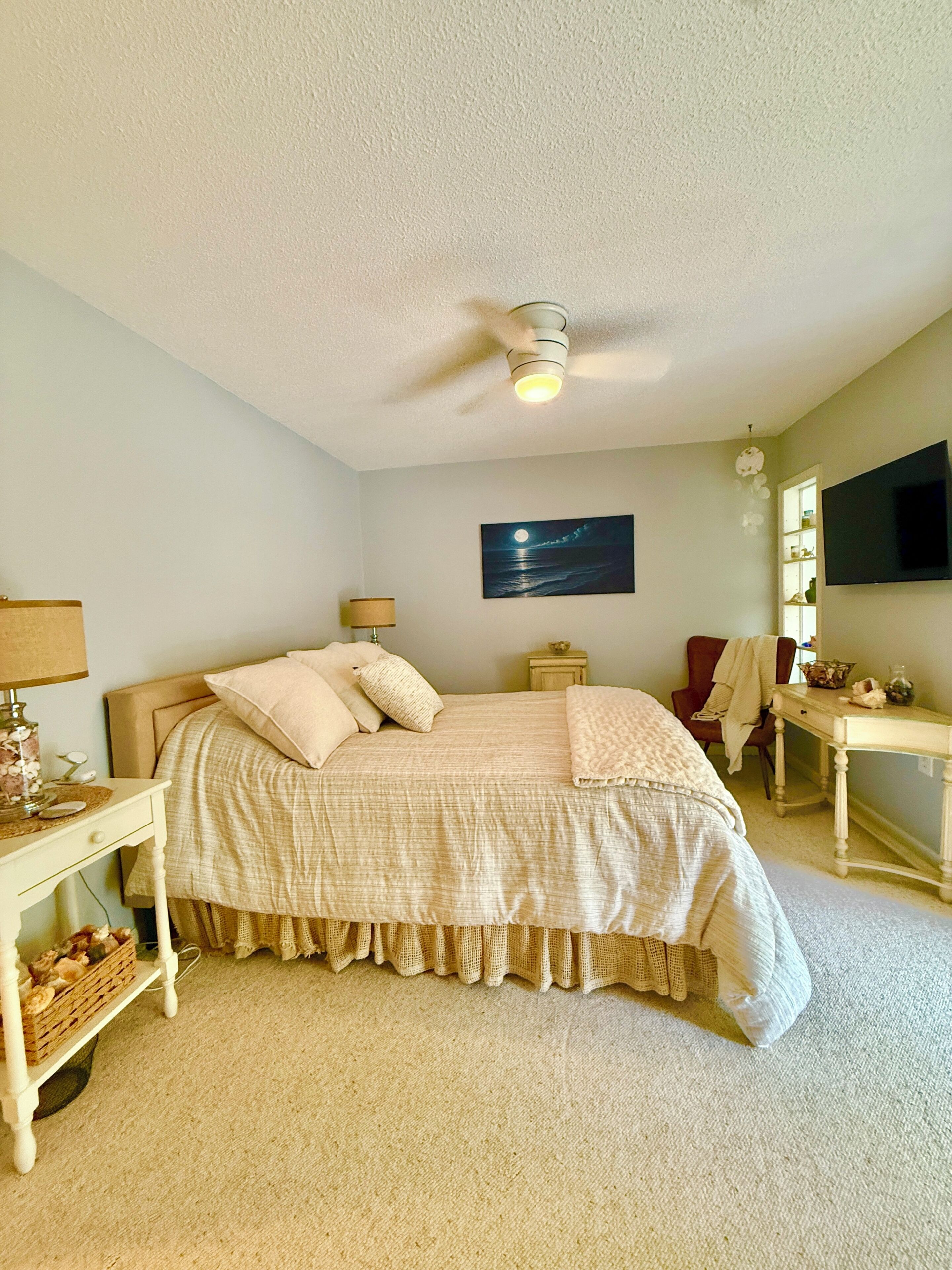Um quarto com uma cama, mesas de cabeceira, abajur, um quadro, televisão e ventilador de teto.