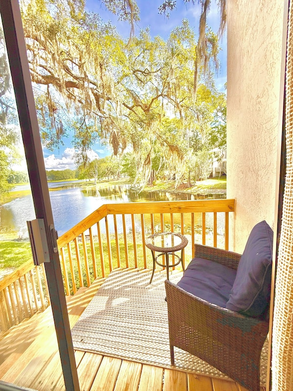 A wooden deck with a wicker chair and a small table, overlooking a body of water and trees.